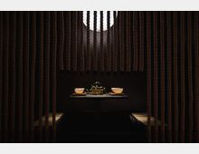 Ein dunkler Raum mit einem niedrigen Tisch in der Mitte, der von Seilvorhängen umgeben ist. Auf dem Tisch stehen zwei Schalen, eine Teekanne und Pflanzenranken, beleuchtet von einer runden, diffusen Lichtquelle im Hintergrund.