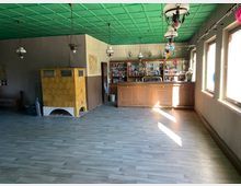 Ein leerer, rustikaler Gastraum mit Holzboden, einer hölzernen Theke im Hintergrund, einer gelben Kachelheizung an der linken Wand und großen Fenstern auf der rechten Seite, die Tageslicht hereinlassen. Die grüne Decke ist mit hängenden Lampen geschmückt.