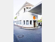 Weißes Gebäude an einer Straßenecke mit einem runden Eingangsbereich und mehreren modernen Fenstern; ein orangefarbenes Schild mit der Aufschrift "ANHALT" über der Eingangstür. Die Umgebung zeigt eine gepflasterte Straße und ein offenes Metalltor als Überdachung vor dem Eingang.