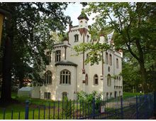Helles, burgartiges Gebäude im neugotischen Stil mit weißen Wänden und dekorativen roten Backstein-Elementen, umgeben von Bäumen und einem Garten. Ein blauer Zaun umgibt das Gelände.
