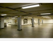 Unterirdischer U-Bahn-Bereich mit gefliesten weißen Wänden, mehreren metallischen Säulen und einer Rolltreppe auf der linken Seite. Die Decke ist flach und von Deckenleuchten beleuchtet, einige schwarz-weiße Wandbilder sind sichtbar.