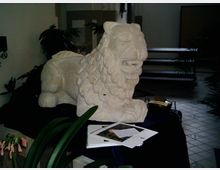 Steinskulptur eines Löwen in einem Innenraum, umgeben von Pflanzen und auf einem dunklen Tuch platziert. Im Vordergrund liegen Dokumente, und der Fliesenboden sowie Möbel im Hintergrund deuten auf eine Ausstellungs- oder Museumsumgebung hin.