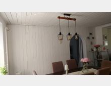 Heller Essbereich mit weißer Holzvertäfelung an Wänden und Decke, einem Esstisch mit Polsterstühlen und einer dreiflammigen Pendelleuchte mit schwarzen Metallkäfig-Schirmen. Im Hintergrund befindet sich ein rundbogiger Durchgang sowie ein Regal mit Deko und Bilderrahmen an der Wand.