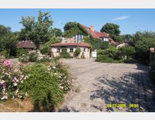Ein ländliches Anwesen mit einem mit Efeu bewachsenen Haus aus Backstein, roten Dachziegeln und weißen Fenstern. Der gepflasterte Hof ist umgeben von blühenden Sträuchern, Bäumen und einer gepflegten Gartenlandschaft.