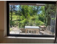 Blick durch ein großes Fenster auf eine Terrasse mit weißem Tisch und mehreren Metallstühlen. Dahinter liegt ein üppig bewachsener Garten mit Bäumen, Sträuchern und einem schmalen Weg.