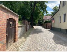 Kopfsteinpflasterstraße in einer ländlichen Umgebung mit altem Mauerwerk, einer Holztür und einem Schild „Weinkeller“ auf der linken Seite. Rechts sind ein helles Wohnhaus und Verkehrsschilder zu sehen, im Hintergrund Bäume unter blauem Himmel.