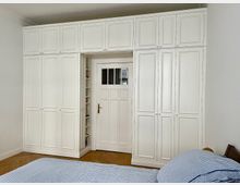 Ein Schlafzimmer mit weißen, deckenhohen Einbauschränken, die eine Tür mit integriertem Glasfenster umrahmen. Im Vordergrund ein Bett mit blauer gestreifter Bettwäsche und Parkettboden.