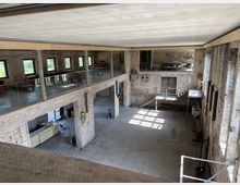 Großer, loftartiger Raum in einem industriellen Gebäude mit freiliegenden Ziegelwänden, einer Galerieebene mit Glasgeländern und großen Fenstern, die Tageslicht hereinlassen. Der Raum ist schlicht eingerichtet und hat Betonböden sowie Stahlgeländer an Treppen und Absätzen.