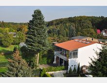 Zweistöckiges weißes Haus mit rotem Ziegeldach und Balkon, umgeben von hohen Nadelbäumen und einem grünen Garten. Im Hintergrund ist ein bewaldeter Hügel und weitere Häuser in ländlicher Umgebung zu sehen.