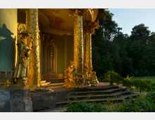 Goldverzierter Pavillon mit reich dekorierten, vergoldeten Säulen und detaillierten Skulpturen von Menschen in asiatisch inspirierten Gewändern; im Hintergrund sind grüne Bäume eines Parks zu sehen.