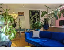 Wohnzimmer mit Holzboden, einem blauen Sofa und zahlreichen Zimmerpflanzen in modernen Töpfen. Im Hintergrund ein Esstisch mit Stühlen, eine Tür sowie urban gestaltete Wanddekorationen, darunter ein großes, Skelett-Motiv an der Wand.