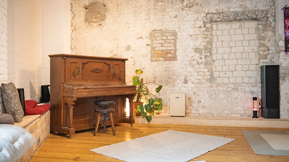 Innenraum im Loft-Stil mit freigelegter, teilweise verputzter Ziegelwand und Holzdielenboden, an der Seite ein altes Klavier mit Hocker. Daneben stehen eine Topfpflanze, kleine Leuchten und ein Sitzpodest mit Kissen; auf dem Boden liegen helle Teppiche.