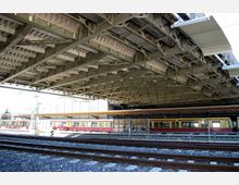 Ein Bahnhof überdacht von einer großen Metallkonstruktion mit freiliegenden Streben. Im Vordergrund verlaufen Bahngleise, während im Hintergrund ein gelb-roter Zug an einem Bahnsteig steht.