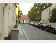 Schmale, gepflasterte Straße mit geparkten Autos auf der rechten Seite und Häusern mit heller Fassade links und rechts. Im Hintergrund steht ein historisches Fachwerkhaus mit rotem Dach, umgeben von Bäumen.