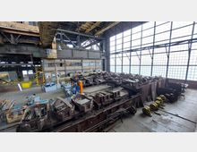 Industriehalle mit hohen Decken und großen Rasterfenstern, die viel Licht einlassen. Im Raum befinden sich massive Metallteile, Arbeitsgeräte und Maschinen, sowie ein Bereich mit Arbeitsstationen und gelben Sicherheitsmarkierungen.