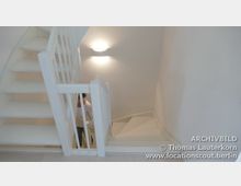 Weiße, lichtdurchflutete Wendeltreppe mit schlichtem Geländer in einem minimalistischen Innenraum; eine Person ist teilweise sichtbar und geht die Treppe hinunter.