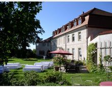 Historisches Gebäude mit hellem Putz und rotem Ziegeldach, umgeben von einem Garten mit Bäumen, Wiesen und Blumenbeeten. Im Vordergrund befinden sich Sitzgelegenheiten, mit weißen Stoffen bedeckte Tische und ein roter Sonnenschirm.