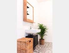 Helles Badezimmer mit einer Wand aus weißen Fliesen, einem Waschbecken auf einem schwarzen Unterschrank mit goldenen Griffen, einem hölzernen Spiegelrahmen darüber, einem geflochtenen Wäschekorb und einer Pflanze im Korb auf gemustertem Fliesenboden.