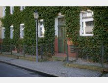 Wohnhausfassade mit efeubewachsenen Ziegelwänden, grüner Haustür mit floralem Glaseinsatz und weißen Fenstern. Im Vordergrund ein schmiedeeiserner Zaun, ein gepflasterter Gehweg und eine Straßenlaterne.