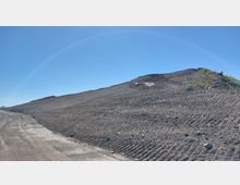 Schotter- und Erdhalde an einer Baustelle unter freiem Himmel, mit leichtem Grünbewuchs auf der rechten Seite. Links ist eine grob geschotterte Straße zu erkennen, die an der Halde entlangführt.