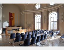 Heller Versammlungssaal mit hohen Rundbogenfenstern, blauen Stühlen in Reihen, einem Rednerpult und einer erhöhten Bühne mit Tischen und Mikrofonen; im Hintergrund eine senkrechte Flagge mit einem Wappen.