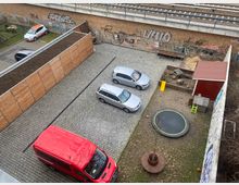 Blick auf einen gepflasterten Parkplatz mit geparkten Autos, neben einem kleinen Spielplatz mit Sandkasten, rotem Spielhaus, gelber Rutsche und einem Trampolin. Im Hintergrund verläuft eine mit Graffiti besprühte Eisenbahnbrücke.
