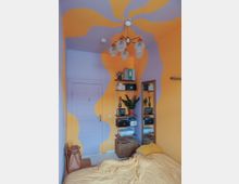 Kleines Schlafzimmer mit gelb-lila geschwungenen Wand- und Deckenmustern, einem hängenden Kronleuchter mit Glaskugeln, einem Regal mit Pflanzen und Ordnern, einem großen Wandspiegel und einem Bett mit gelber Bettdecke. Im Hintergrund ist eine weiße Tür sichtbar.