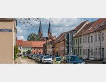 Straßenszene mit Reihenhäusern in hellen Farben und roten Ziegeldächern, geparkte Autos entlang der Straße; im Hintergrund ragen die zwei spitzen Türme einer Backsteinkirche in den Himmel. Schild mit der Aufschrift "Rudolf-Breitscheid-Str." sichtbar im Vordergrund.