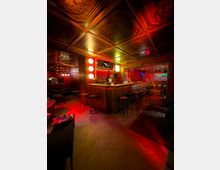 Innenraum einer Bar mit langer Holztheke und hohen Barhockern, rot-gelb beleuchtet mit dekorativen Wandlampen und einem Fernseher über dem Tresen. Im Hintergrund sind Lounge-Sitzbereiche und eine gemusterte, kassettierte Decke mit Ornamentelementen zu sehen.