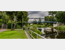 Metallbrücke über einen kleinen Fluss mit grünen Uferbereichen, dichtem Baumbewuchs und einem Gehweg mit Treppe. Im Wasser ist ein weißes Boot zu sehen, und eine Person steht am Ufer.