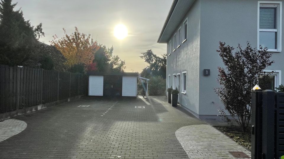 Wohngebiet mit einem zweistöckigen hellgrauen Gebäude auf der rechten Seite, gepflastertem Hof und zwei Garagen am Ende des Weges. Im Hintergrund ist ein Baum mit Herbstlaub zu sehen, und die Abendsonne scheint durch einen leicht bewölkten Himmel.