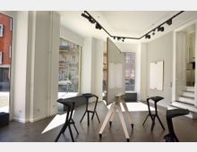 Heller, moderner Seminarraum mit bodentiefen Fenstern, mobilen Whiteboards, schwarzen Hockern und einer Treppe im Hintergrund. Der Raum hat weiße Wände und Deckenleuchten in Schienen.