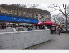 Eingang zur U-Bahn-Station "Rathaus Steglitz" mit einem blauen Schild, umgeben von einem Wochenmarkt mit roten und grünen Verkaufszelten. Im Hintergrund befinden sich Bäume ohne Laub sowie ein mehrstöckiges Wohn- und Geschäftshaus.