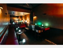 Schmaler, gedämpft beleuchteter Lounge- oder Barraum mit gepolsterten Lederbänken entlang der Wände, kleinen runden Tischen und niedrigen Tischlampen. Rechts stehen hohe Barhocker an einem Wandtisch mit farbigem Licht, im Hintergrund sind Spiegel und weitere Sitzplätze zu sehen.