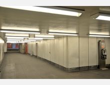 Unterführung mit hell beleuchteten, weißen Deckenplatten und neutralen, grauen Wänden. Links führt eine Treppe nach oben, während rechts ein öffentlicher Münztelefonautomat steht.