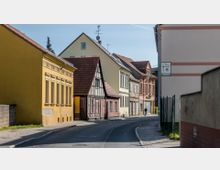 Straßenszene in einer Kleinstadt mit eng beieinanderstehenden Häusern im historischen Stil, darunter ein gelbes Gebäude mit klassischer Fassadengestaltung und ein Fachwerkhaus. Die schmale, leicht kurvige Straße ist asphaltiert und von Gehwegen gesäumt.