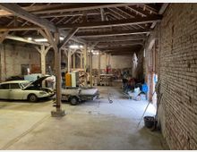 Innenansicht einer großen Scheune mit freiliegendem Dachgebälk und unverputzten Ziegelwänden. Im Raum stehen ein Oldtimer mit geöffneter Motorhaube, ein kleines Boot auf einem Anhänger sowie verschiedene Werkzeuge, Holzstapel und Baustellenmaterialien.