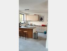 Moderne Küche mit heller Wand und minimalistischen Schränken in weißem Design. Zentral steht eine Kücheninsel mit Holzverkleidung und zwei Hockern, ergänzt durch ein großes Fenster, einen roten Toaster und eine Wanddekoration.