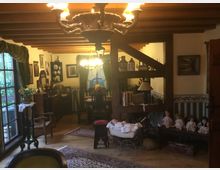 Gemütliches Wohnzimmer im rustikalen Stil mit Holzbalkendecke und antiken Möbeln. Im Raum befinden sich alte Puppen, ein Kinderwagen, Teppiche, sowie dekorative Objekte und Bilder an den Wänden, mit einem großen Fenster, das Tageslicht hereinlässt.