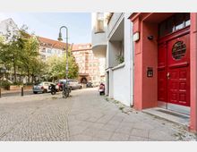 Straßenszene in einer Wohngegend mit Kopfsteinpflaster, einem roten Hauseingang im Vordergrund und mehreren geparkten Autos und Fahrrädern entlang der Straße. Im Hintergrund stehen mehrstöckige Gebäude mit verzierten Fassaden und roten Dächer, sowie Bäume, die den Straßenrand säumen.