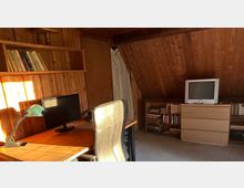 Ein Arbeitszimmer mit Holzvertäfelung und Dachschräge. Zu sehen sind ein Schreibtisch mit Monitor, Lampe und Lederstuhl, ein Regal mit Büchern an der Wand sowie eine Kommode mit einem kleinen Röhrenfernseher darauf.