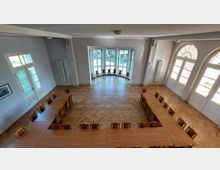 Großer, heller Veranstaltungsraum mit Parkettboden, rechteckigen Tischen in U-Form und Holzstühlen. Der Raum hat hohe Decken, große Fensterfronten und weiße Doppeltüren, die reichlich Tageslicht hereinlassen.