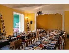 Innenraum eines Veranstaltungs- oder Speiseraums mit gelben Wänden, langen gedeckten Tafeln und vielen gemischten Stühlen. Im Hintergrund sind eine große bogenförmige Nische, ein Deckenprojektor und eine offene Tür zu einem angrenzenden türkisfarbenen Raum zu sehen.