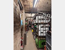 Ein schmaler Lagerraum mit Backsteinwänden und gewölbter Decke. Regale voller großer Buchstaben, bunter Objekte und Kisten säumen den Gang, der von Neonröhren beleuchtet wird.