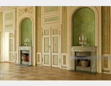 Klassischer Innenraum mit grüner Wandgestaltung und Ornamentverzierungen, zwei dekorativen Kaminen mit Marmorrahmen sowie einem Doppelflügeltürensemble in der Mitte. Der Raum hat Parkettboden und eine historische, kunstvolle Gestaltung.