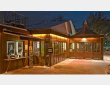 Ein hölzerner Kassenpavillon bei Nacht mit beleuchteten Fenstern und einem Spitzdach auf der rechten Seite. Der Boden ist schneebedeckt, und im Hintergrund sind Bäume und ein dunkler Himmel zu sehen.