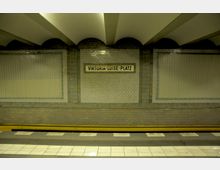 U-Bahn-Station mit einem Wandpaneel aus weißen Fliesen, auf dem der Stationsname "Viktoria-Luise-Platz" zu lesen ist. Der Bahnsteig ist mit gelben Sicherheitsleisten und einem Kachelboden gestaltet.