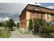 Ein zweistöckiges Gebäude mit roter Fassade und Ziegeldach, beschriftet mit „Zum alten Schloß“, mit einem kleinen Biergarten und Sonnenschirmen im Vordergrund. Daneben führt ein gepflasterter Weg durch eine grüne Landschaft mit Bäumen und eingezäunten Wiesen.