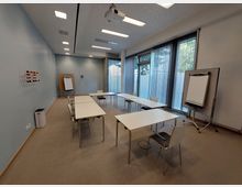 Moderner Seminarraum mit blauen Wänden und großen Fenstern, die durch vertikale Jalousien abgedeckt sind. Der Raum ist mit weißen Tischen und Stühlen ausgestattet, einem Flipchart, einer Pinnwand sowie einem Deckenprojektor.