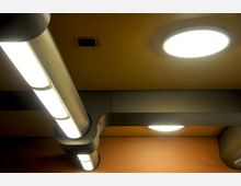 Decke eines Innenraums mit modernen, vertikal und horizontal verlaufenden Leuchtstofflampen, ergänzt durch eine runde Deckenleuchte. Die Deckenfläche ist in warmen Gelb- und Orangetönen gestaltet, mit metallischen Elementen.